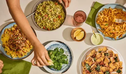 Table spread of Pulled Pork BBQ Mac &amp; Cheese, Basil Pesto Cavatappi, Lemon Parmesan Broccoli, Pasta Fresca, Cajun Shrimp Fettuccinne. Hand squeezing lemon over Lemon Parmesan Broccoli.